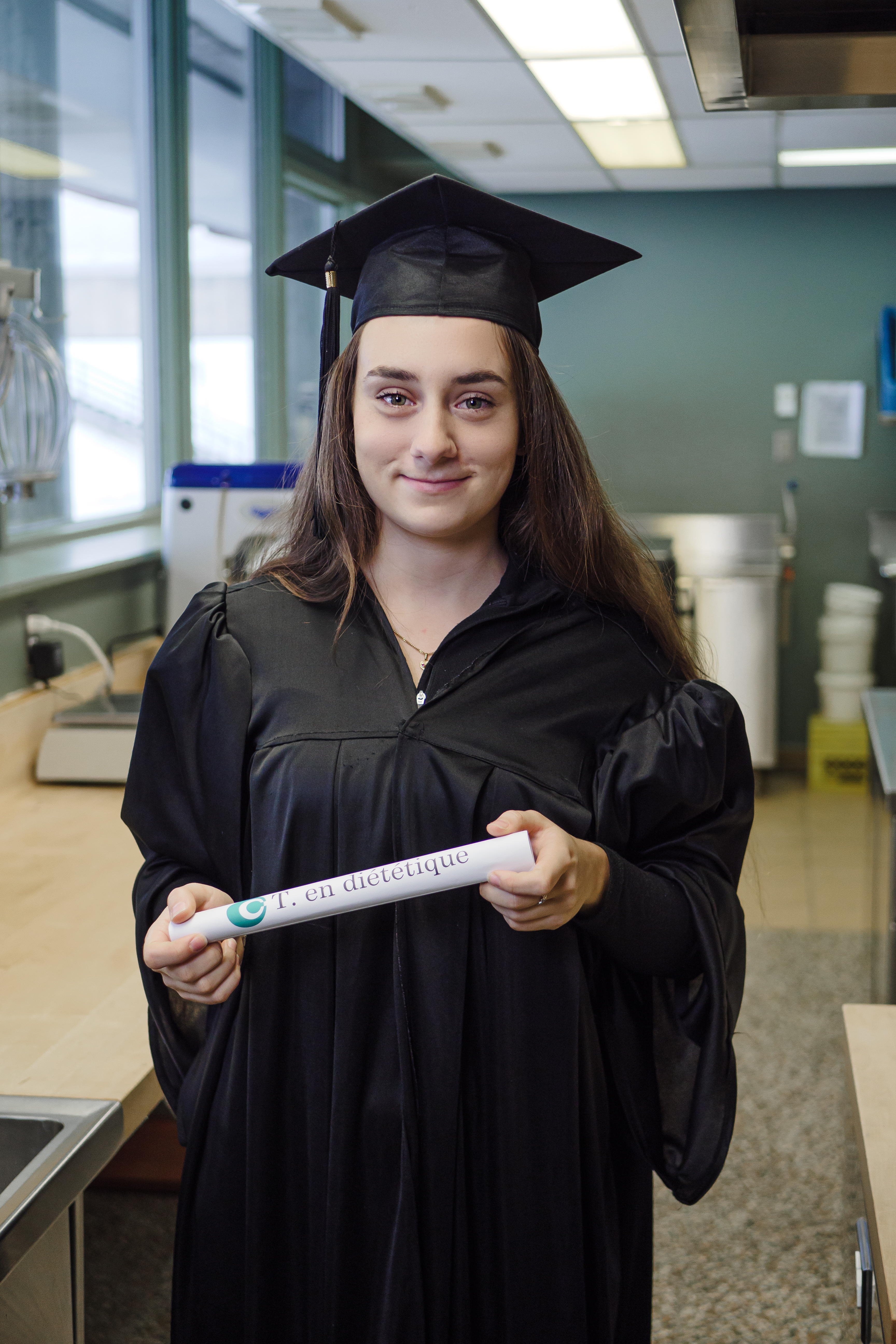 Je suis en robe de remise de diplôme. Dans mes mains, je tiens mon diplôme. En arrière plan, nous apercevons une cuisine.
